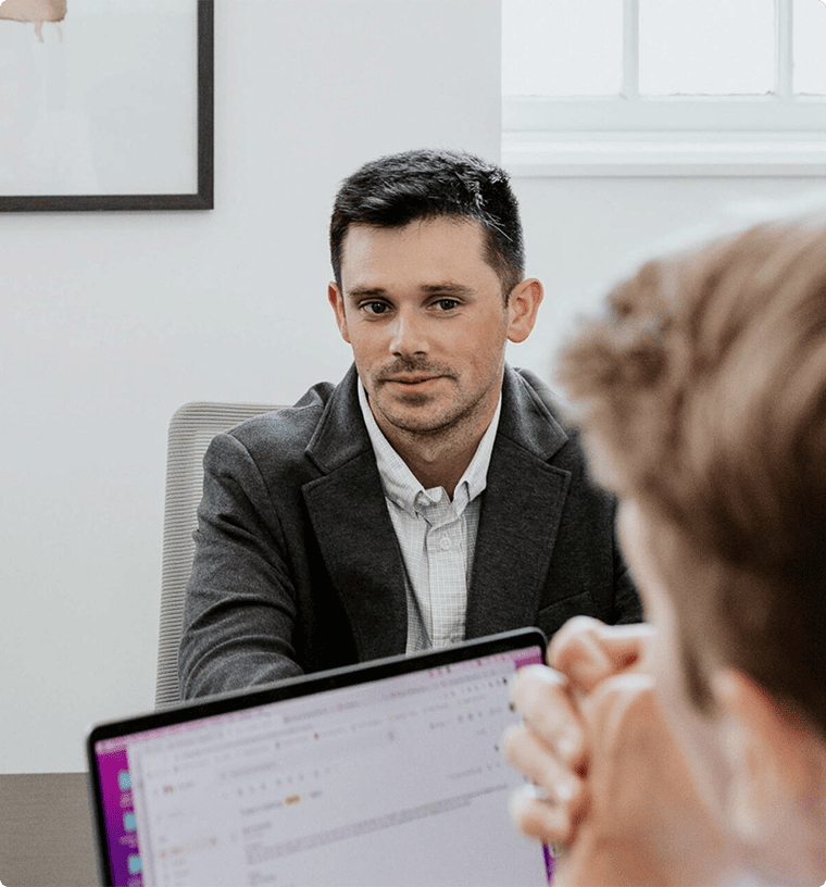 A man in a suit sits at a table in an office setting, looking towards another person with a laptop, whose back is to the camera.