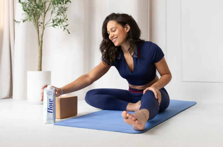 Person in a navy athletic outfit sits cross-legged on a blue yoga mat, reaching for a can with a yoga block nearby. An indoor plant is in the background.