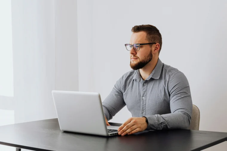 Man with glasses working on a laptop at a black table in a bright room.