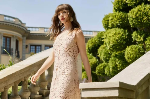 A woman in a lace dress walks down stone steps outside, with a large building and greenery in the background.