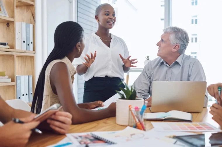 A diverse group of people in a meeting room; a woman stands and speaks while others, seated, listen attentively. A laptop, documents, and stationery are on the table.
