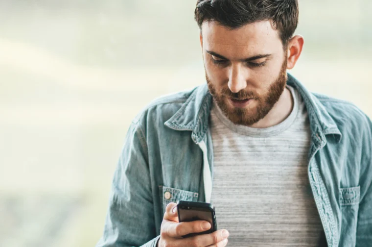 Man with a beard looks at a smartphone while wearing a denim shirt over a gray t-shirt.