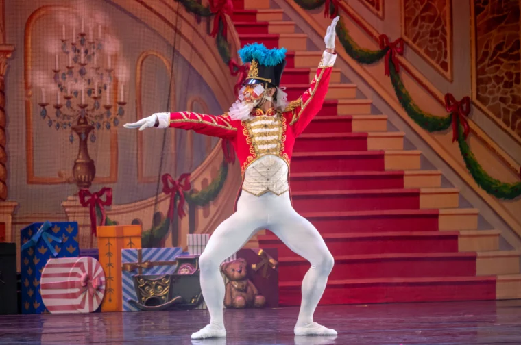 Ballet dancer in a colorful Nutcracker costume performs on stage with festive decorations, including gifts and a staircase in the background.