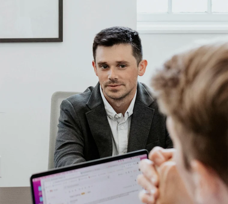 A man in a suit sits at a table, looking towards another person who is partially visible. A laptop is open in the foreground.