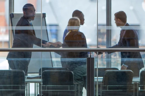 Four people in business attire are in a meeting room, two are shaking hands, and the others are nearby. A flip chart is in the background, and the scene is viewed through glass windows.