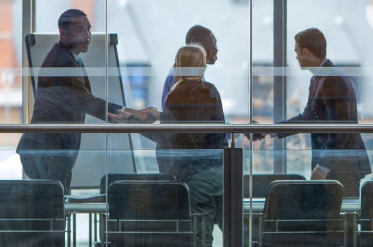 Four people in business attire are in a meeting room, two are shaking hands, and the others are nearby. A flip chart is in the background, and the scene is viewed through glass windows.