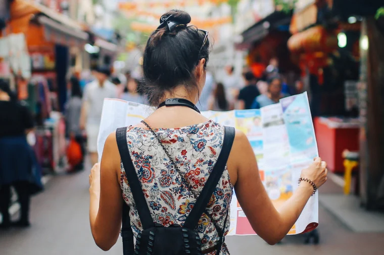 A woman stands in a bustling market street, holding a map. She has a backpack and is wearing a floral top, with her hair in a bun. The market around her is busy and colorful.