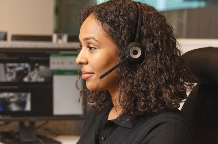 A woman with curly hair is wearing a headset and sitting at a desk with computer monitors.
