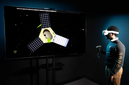 A person wearing a VR headset interacts with a virtual satellite displayed on a large screen.