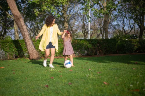 A woman and a child play with a soccer ball on a grassy field surrounded by trees.