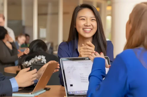 People sitting at a table, engaged in a discussion. A woman is smiling, holding her hands together. Laptops are open in front of them.