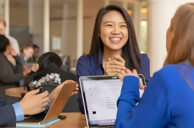 People sitting at a table, engaged in a discussion. A woman is smiling, holding her hands together. Laptops are open in front of them.