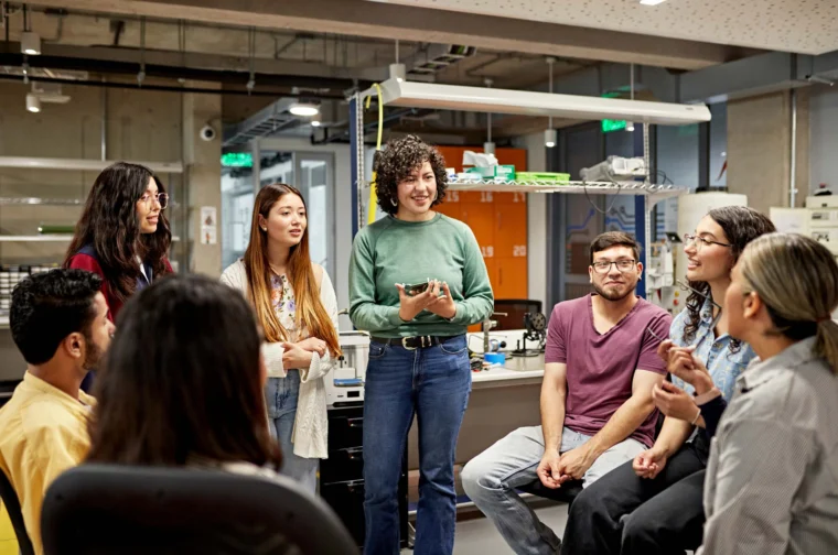 A group of people engage in a discussion in a modern workspace with electronic equipment in the background.