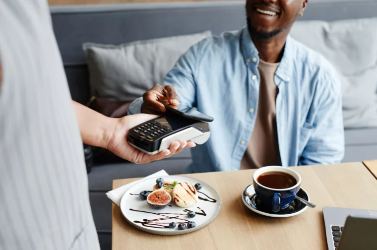 A person uses a smartphone for contactless payment at a café. A plate with dessert and a cup of coffee are on the table. Another person holds the card reader.
