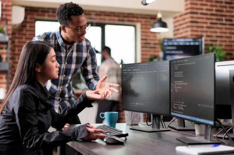 Two people are working at a desk with dual monitors displaying code. One is seated, typing, while the other stands, gesturing towards the screen in an office setting.
