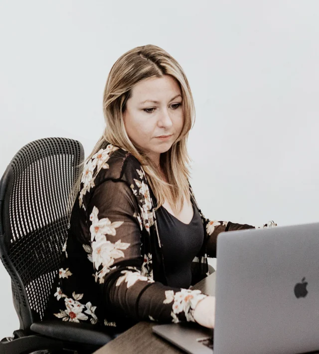 Person seated at a desk, working on a laptop, wearing a black floral top.