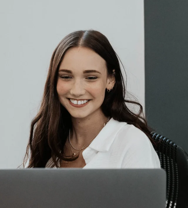 A person with long brown hair and a white shirt is smiling while looking at a laptop.