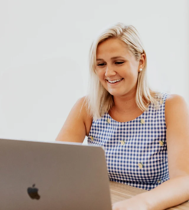 A person with blonde hair smiles while using a laptop. They are wearing a blue and white checkered top with small yellow patterns.