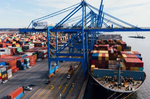Cargo ship docked at a busy port with blue cranes loading colorful shipping containers onto the vessel, surrounded by stacked containers and vehicles on the dock.