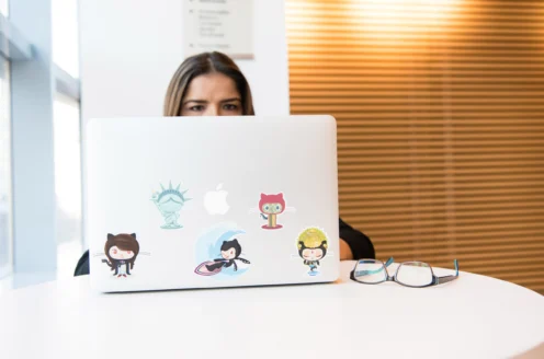 Person sitting at a table using a laptop covered in various stickers, with eyeglasses placed nearby.