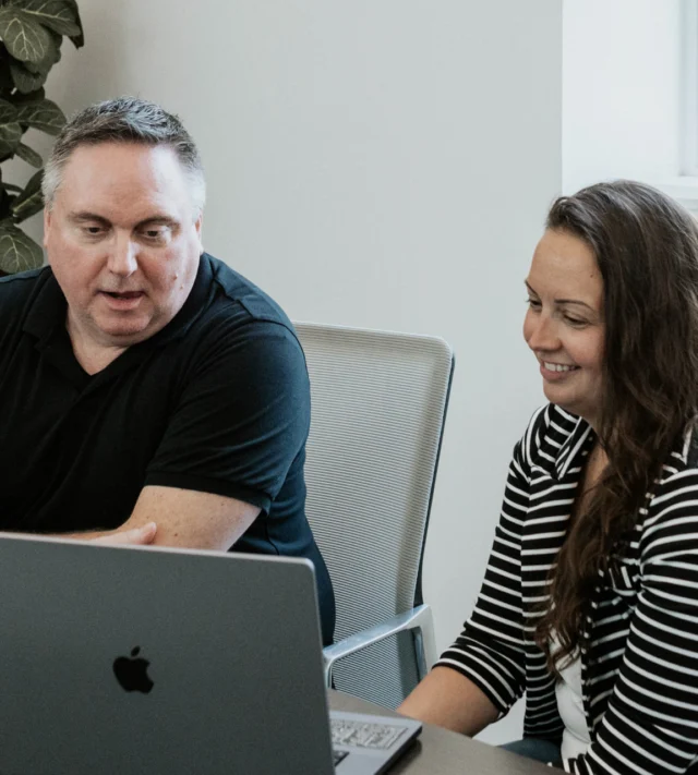 Two people sit at a table, looking at a laptop. The man points at the screen while speaking, and the woman smiles, appearing engaged in the discussion.