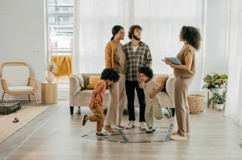 A family stands in a living room while a person with a tablet speaks to them. A child jumps in the foreground.