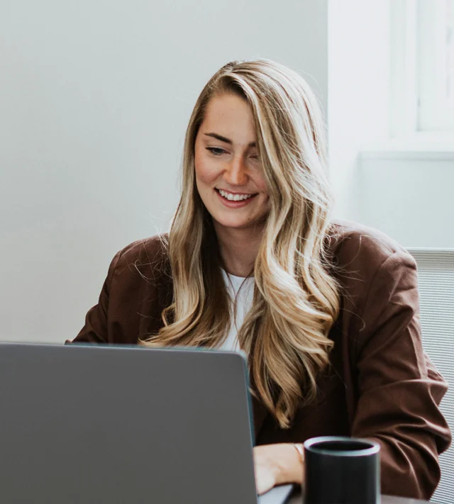 A person with long hair smiling while working on a laptop, with a mug nearby.