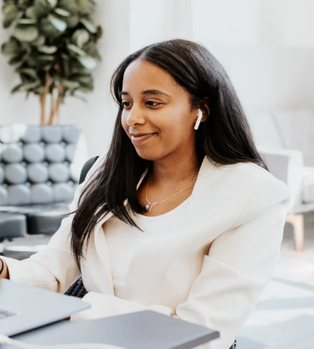 A woman in a white blazer sits at a desk wearing wireless earbuds, looking at a laptop. There's a plant and a chair in the background.