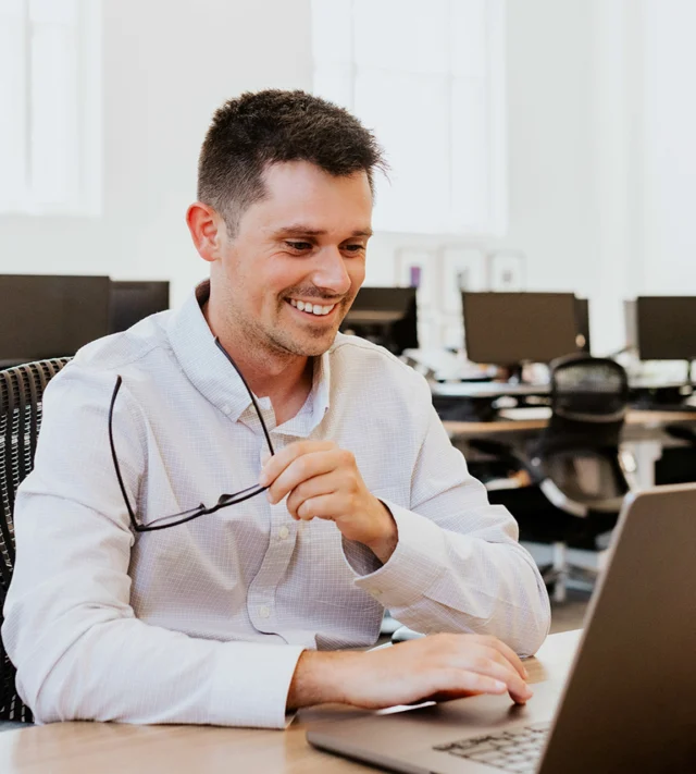 Person in a white shirt sits at a desk holding glasses, smiling while using a laptop in an office setting.