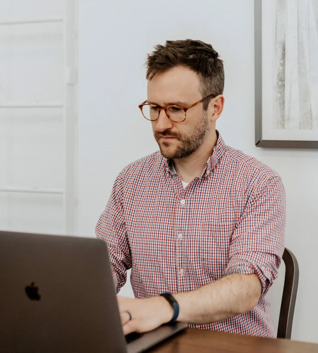 Man wearing glasses and a checkered shirt sits at a table, focused on typing on a laptop.