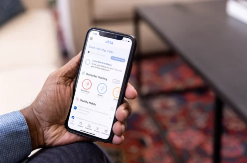 Person holding a smartphone displaying a health app interface with options for biomarker tracking and healthy habits, on a colorful carpeted background.