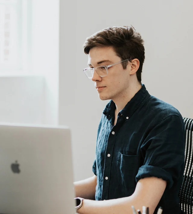 Person with glasses and short hair working on a laptop at a desk in a bright room.