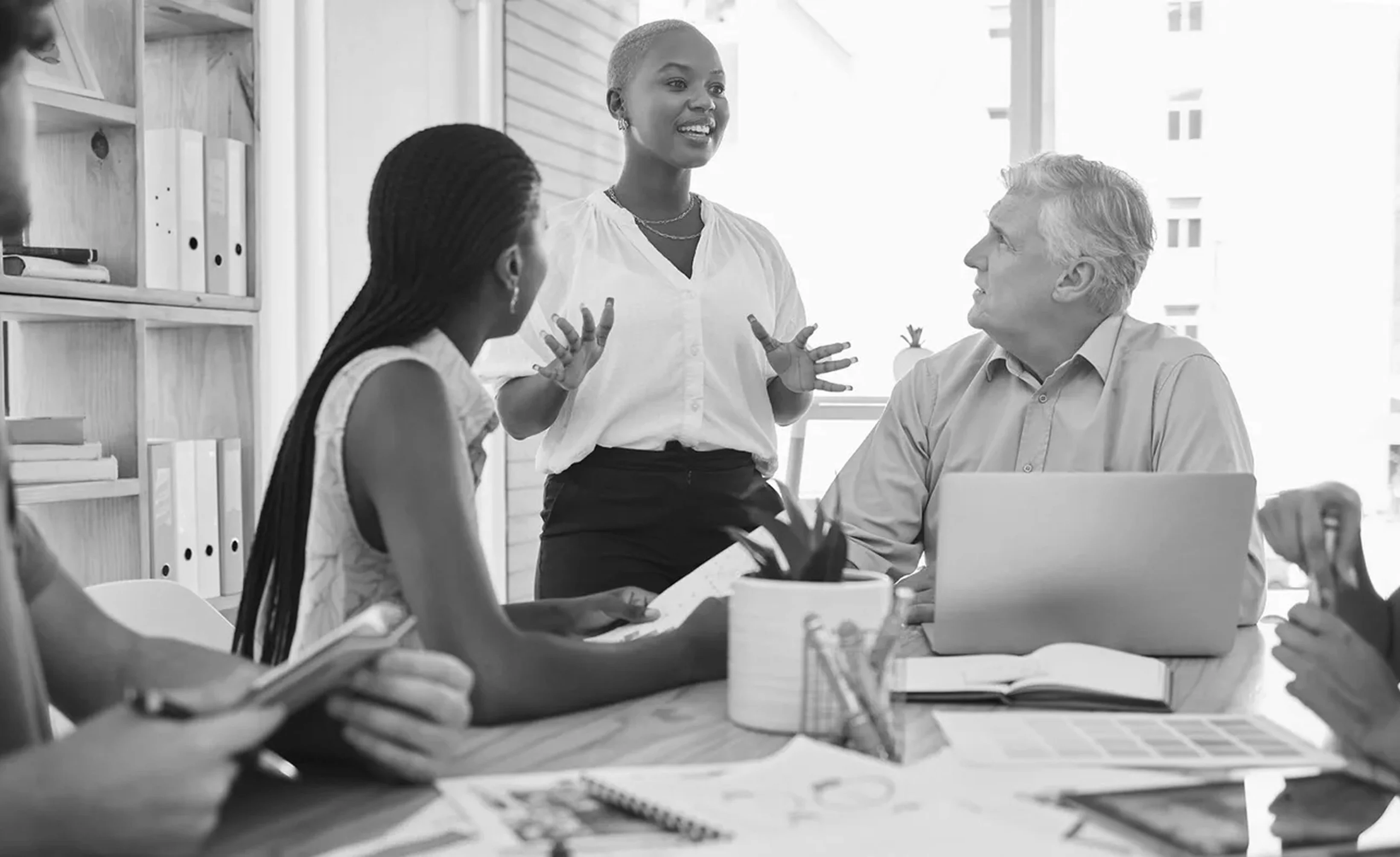 A person stands speaking to two seated individuals at a table with laptops and documents in a bright office setting.